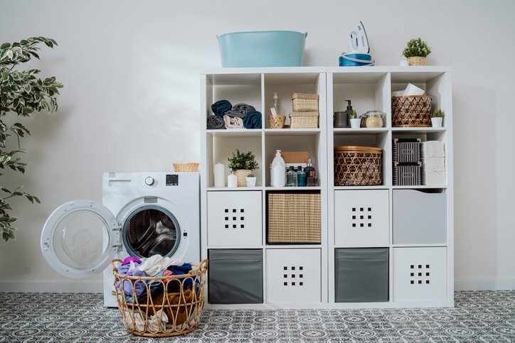 View of home laundry room, dresser with softeners, powder, towels, open washing machine with empty drum, next to wicker basket with colorful wash items