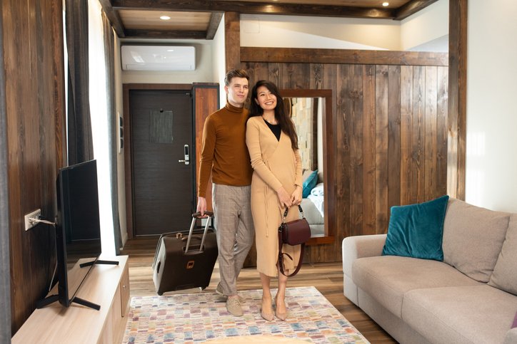 Young Asian woman in elegant beige dress looking at interior of hotel room while her husband or boyfriend with suitcase standing near by
